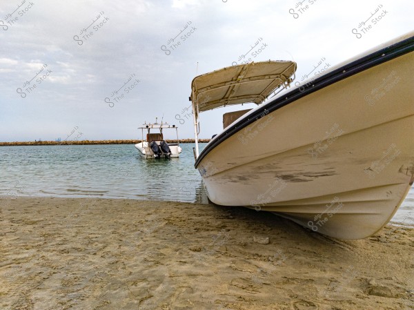 A large boat in the foreground on a sandy beach with a canopy, with another boat in the background floating in calm waters. The sky is cloudy in the distant horizon, with a breakwater visible in the distance.