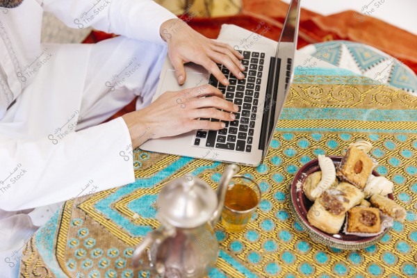 An image of a person wearing a traditional white thobe sitting on a vibrant, decorative carpet with colors such as blue and gold, using a laptop. On the table, there is a metal teapot with a small glass of tea and a plate of assorted traditional pastries.