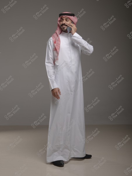 Image of a man wearing a white thobe and a red and white checkered ghutra with a black agal. He is standing, smiling, and talking on a mobile phone. The background is a neutral grey color.