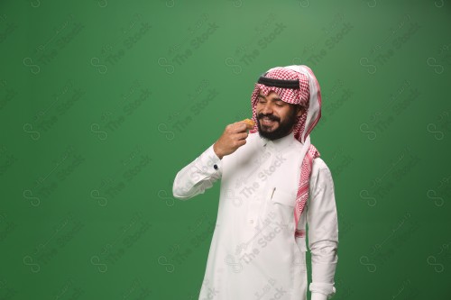 شاب سعودي على خلفية خضراء يرتدي الثوب و الشماغ، يقوم بعمل حركات بيده، يأكل الكعكة ، احتفالات العيد