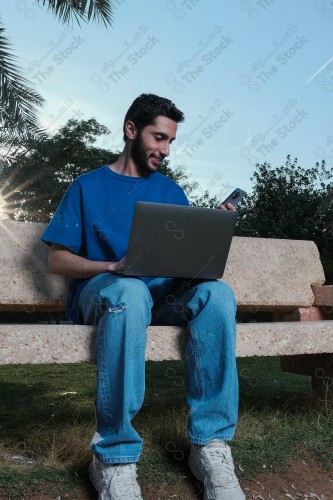 A Saudi man sits on a sofa in a public park and uses a mobile device during the day.