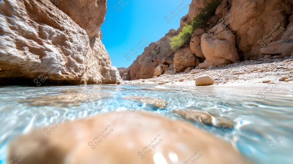 A natural scene of a shallow water stream flowing between rocks in an arid mountainous region. The clear water reflects light under the bright sunlight, surrounded by beige-colored rocks and mountains, with a few trees visible in the background.