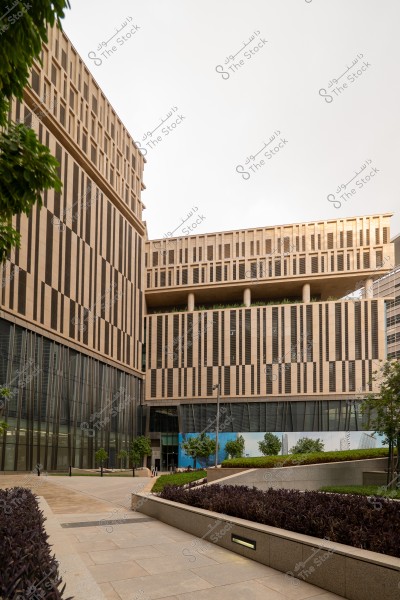 A modern building with a distinctive architectural design located in an urban area. The building features large glass windows and vertical lines in harmonious beige and brown colors. In the foreground, there is an open space with paved walkways, carefully planted shrubs, and greenery. The sky is overcast, casting a muted tone in the background.
