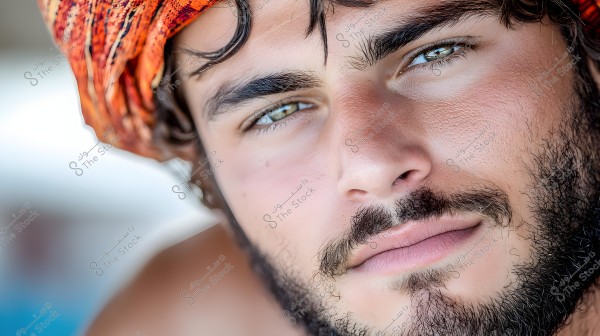 A portrait of a man wearing a colorful orange headscarf with pronounced facial features and a light beard. The background is blurred, suggesting possibly a bright outdoor setting.