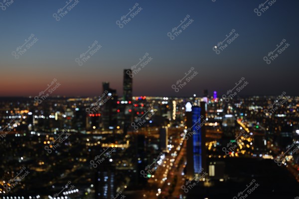 The image shows an aerial view of a vibrant city in the evening or night. The city lights twinkle, giving an impression of traffic and activity in the tall buildings. The sky is dark with a slight twilight on the horizon, and there is a prominent blue-lit tower in the foreground.