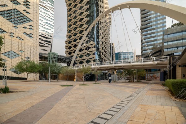 Image of a modern urban street featuring a suspension bridge and several high-rise buildings with unique architectural designs. The buildings have intricate geometric patterns, with some having glass facades. Green plants are dispersed along the street, adding a natural touch to the scene.