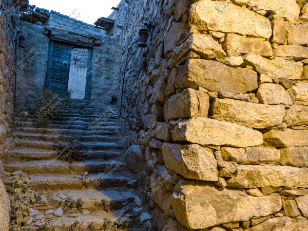 The image shows an old stone alley with steps leading up to a blue wooden door at the end of the passageway. The surrounding stone walls exhibit signs of aging with dust accumulation and plants growing between the stones, and an old lantern is mounted on the right wall.