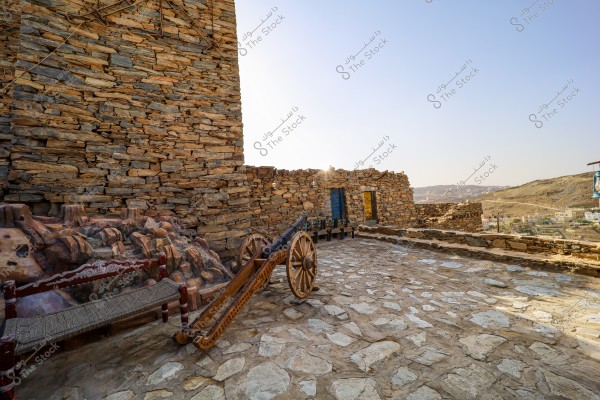 View of a traditional stone courtyard in a mountainous area, featuring an old stone wall and a wooden cannon with wooden wheels. Traditional wooden benches are placed alongside the wall, and colorful lamps are hanging on the wall. In the background, the landscape of mountains and houses is visible.