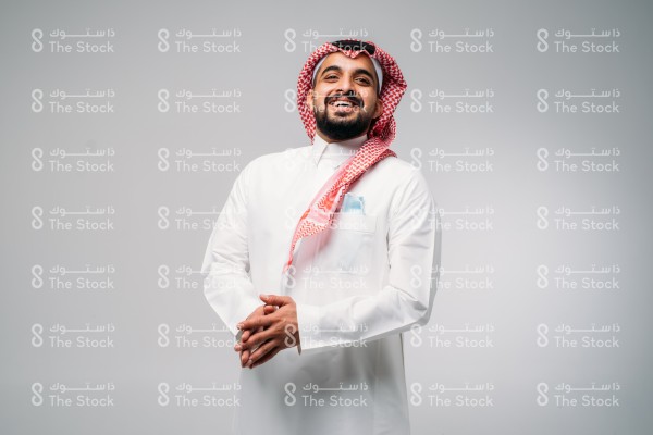 بورتريه رجل سعودي يرتدي الزي السعودي التقليدي يرفع رأسه مبتسمًا بينما يشبك يديه و تظهر كمامته بالجيب الأمامي