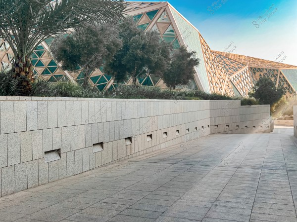 The image shows a gray paved walkway flanked by tall stone walls, with palm trees and other trees in the background. In the distance, there are modern architectural structures with distinctive triangular glass and metal frame designs. The sky is clear and blue.