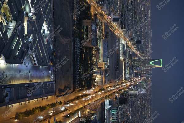 منظر العاصمة السعودية الرياض مساءً من ارتفاع عالٍ, حيث يظهر أبراج حديثة مضاءة بشكل لافت, بينها برج المملكة الذي يغمره الضوء الأخضر. تظهر الشوارع مضاءة بأضواء السيارات والمباني تتناثر على نطاق واسع.