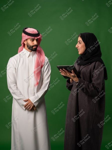 يتفاعل رجل وامرأة سعوديان يرتديان الزي التقليدي في استوديو ذو خلفية خضراء, حيث تحمل المرأة جهازًا لوحيًا.