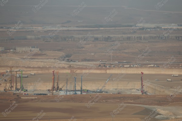 The image depicts a large construction site in an open desert area. Various colorful cranes and drilling rigs are positioned in the center of the excavated site, with trucks and vehicles moving around. In the background, several distant buildings and structures are visible on the horizon, with sprawling sandy and rocky hills. The sky is slightly hazy, suggesting a morning or dusty atmosphere.
