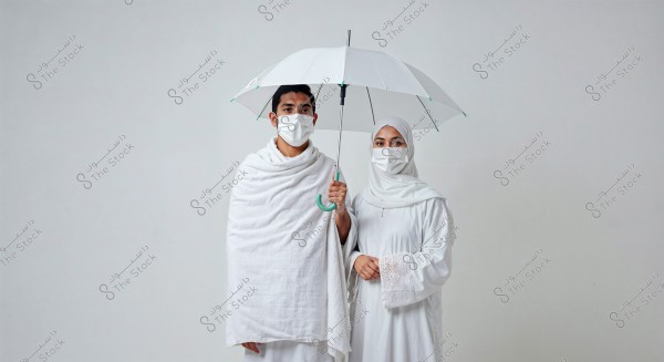 An image of two individuals wearing white Ihram garments, standing under a white umbrella. The first person is dressed in an Izar and Rida, while the second person is wearing a hijab and a long white dress. Both are wearing white face masks.