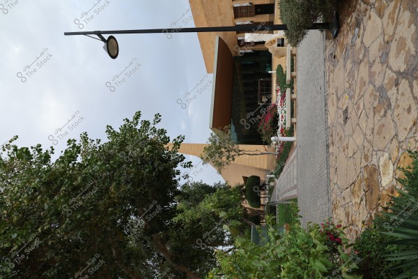 The image shows a modern building with an artistic design located at the end of a stone-paved path surrounded by trees and green plants. The entrance features glass and modern ceilings with colorful flowers planted in the surrounding gardens. There is a tall streetlight on the left and a paved walkway with varied colors and textures.