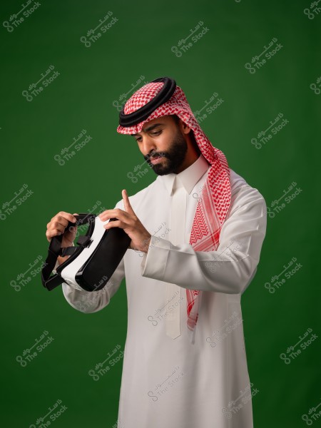 رجل يرتدي الثوب الخليجي التقليدي ويفحص نظارة الواقع الافتراضي أمام خلفية خضراء.