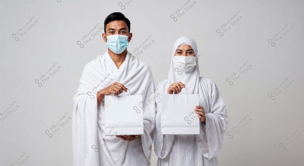 A man and a woman wearing ihram clothing hold white bags. The man is dressed in the traditional white ihram garment, and the woman is wearing a white hijab. Both are wearing medical masks. The background is white and neutral.