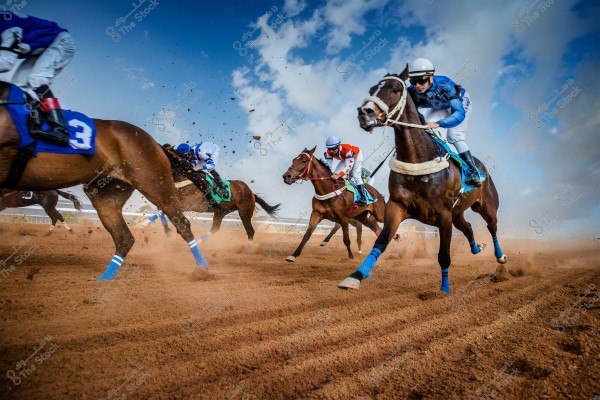 A dynamic scene of an exhilarating horse race featuring several horses and jockeys wearing colorful attire, including blue, white, and red. They are all racing on a sandy track under a blue sky with scattered clouds. The dust trailing behind the horses vividly depicts motion and speed.