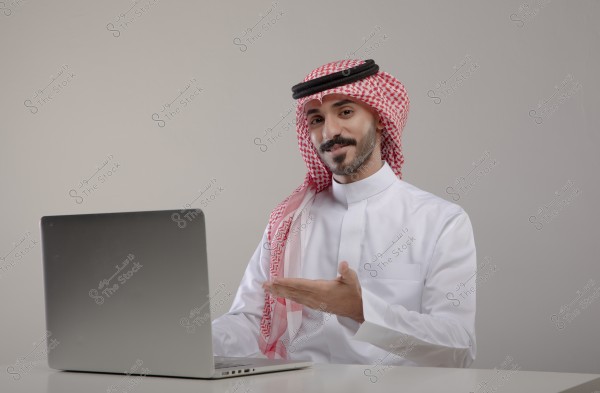 A portrait of a man wearing traditional Saudi attire, consisting of a white thobe, a red checkered keffiyeh, and a black agal, sitting in front of a laptop. The photo appears to be taken indoors with good lighting and a simple background.