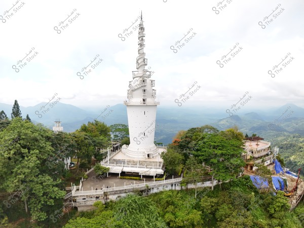 The image shows a tall white tower with a circular ornate design, surrounded by lush green gardens and stunning mountainous nature. The sky is cloudy in the background, and the surrounding trees give the area vibrant green shades.