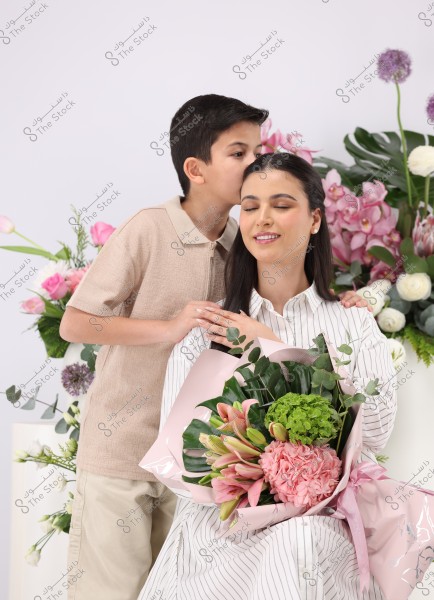 The image shows a woman in a striped shirt holding a bouquet of pink and green flowers, sitting with a smile and her eyes closed. Beside her stands a child in a beige shirt gently kissing her on the head. Their background features beautiful floral arrangements in shades of pink, white, and green.