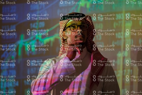 شاب سعودي يرتدي الزي السعودي التقليدي يتابع سوق الأسهم ويظهر انعكاس أضواء الشاشة عليه بينما يضع يده على دقنة يفكر, رجل يتابع سوق المال