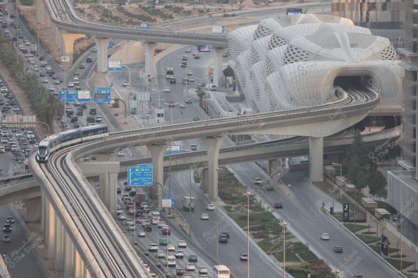 Image of a modern city featuring a multi-level road intersection with cars and trains in motion. A building with a modern architectural design is visible, with a dedicated rail bridge above the road. Trees and plants line the sides of the roads, and traffic signs are present for organization.