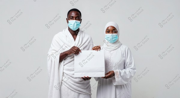 A portrait of a man and a woman standing against a white background, both wearing medical masks. The man is dressed in a white Ihram, while the woman is wearing a white abaya and hijab. The couple is holding a white shopping bag together.
