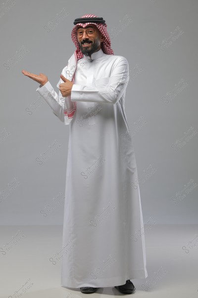 صورة لرجل يرتدي ثوباً أبيض وغترة حمراء وبيضاء, ويشير بيديه بتعبير مرح. الخلفية رمادية بسيطة والمشهد يوحي بالثقة والود.