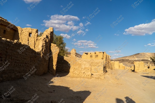 صورة لقرية أو موقع أثري قديم في منطقة صحراوية. يظهر في الصورة أطلال مباني طينية متآكلة, وجدرانها بالية تحت سماء زرقاء صافية تحتوي على بعض السحب المتفرقة. توجد خلف الجدران بعض الأشجار, وتُرى تلال في الأفق.