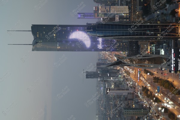 A nighttime photo of Riyadh, Saudi Arabia, featuring a tall, illuminated tower with purple and white lights and distinctive architectural features. The surrounding streets are busy with cars, and the buildings glitter with lights in modern designs. The sky has a soft night blue hue, with light spreading across the horizon.