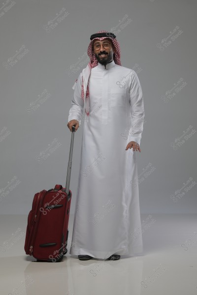 Portrait of a man wearing traditional Saudi attire, including a white thobe, a red and white checkered ghutra, and a black agal. He is holding a red suitcase beside him and appears to be smiling, standing in a studio with a neat and polished appearance.