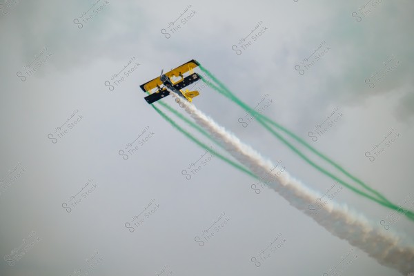 An aerobatic plane flying in the blue sky, releasing white and green smoke forming two parallel trails extending backward. The plane has yellow and blue colors and features a design tailored for air performances.