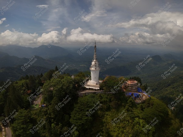 An aerial view of a tall white tower surrounded by dense green forest atop a mountain. A range of mountains can be seen in the distance under a blue sky with white clouds. Several small buildings are visible next to the tower, partially covered with blue tarps.