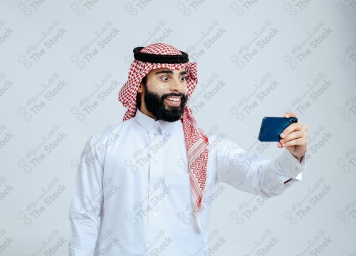 رجل سعودي يرتدي الزي الرسمي التقليدي على خلفية بيضاء, تصفح الهاتف المحمول, دردشه ومحادثات, التواصل مع الأصدقاء, مكالمات فيديو, القراءه من الهاتف المحمول, قضاء وقت ممتع وهادئ, تسليه وترفيه.