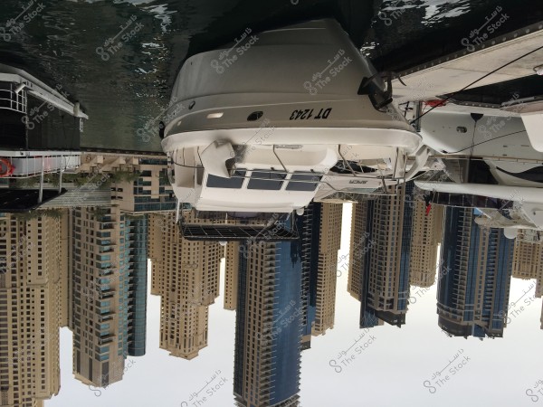 The image shows a white yacht docked at a marina, with calm waters reflecting the tall buildings in the background. The buildings are modern and have a contemporary architectural style, and the gray sky indicates an overcast day. The yacht has a registration number on the side and is securely moored to the dock.