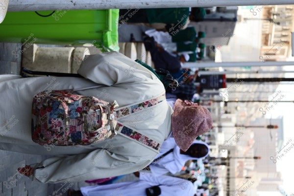 The image shows a person wearing a pink hat and a light-colored jacket with a colorful backpack. They appear to be walking on a busy street in a city, with a group of people in green uniforms in the background, along with others wearing traditional white clothing. Green trash bins are on the side of the road.