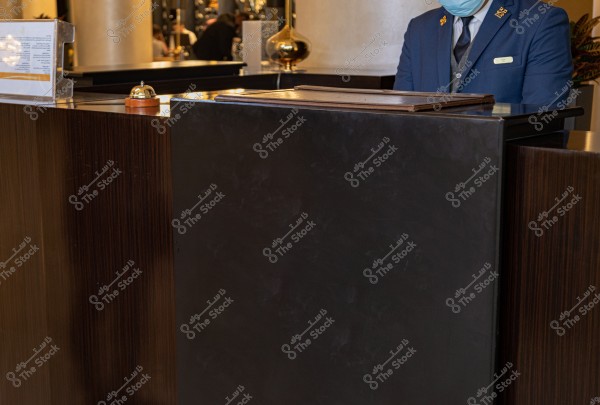 يُظهر الصورة مكتب استقبال في بهو فندق, حيث يقف شخص يرتدي زيًا رسميًا باللون الأزرق وربطة عنق, بالإضافة إلى قناع طبي يغطي وجهه. يتواجد جرس صغير على الطاولة بجانب ملفات ورقية بنهايتها. توجد خلفية مليئة بالزخارف والنباتات.