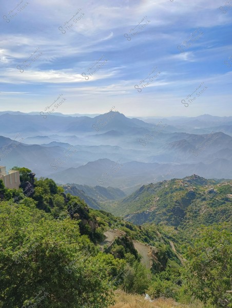 A scenic view of a lush mountainous area with clouds covering the distant peaks. Some houses are scattered on the hills, and dense green vegetation covers the slopes. The sky is blue with light white clouds.