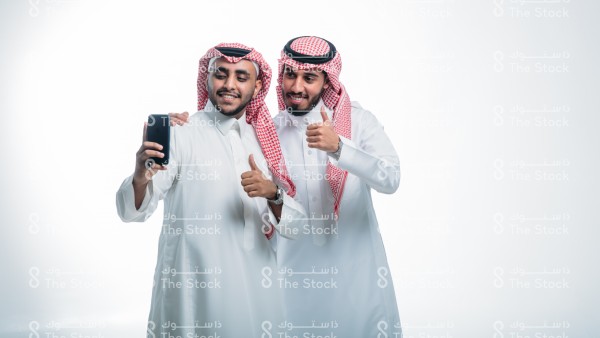 بورتريه صديقان سعوديان يرتديان الثوب ويقفان أمام خلفية بيضاء،  يلتقطان صور سيلفي ، توثيق لحظة سعيدة ،استخدام وسائل التواصل الاجتماعي ، صور تذكارية