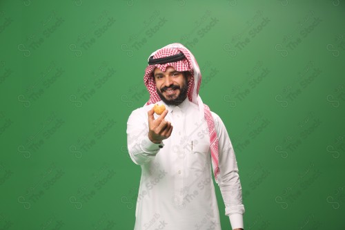 شاب سعودي على خلفية خضراء يرتدي الثوب و الشماغ، يقوم بعمل حركات بيده، يأكل الكعكة ، احتفالات العيد