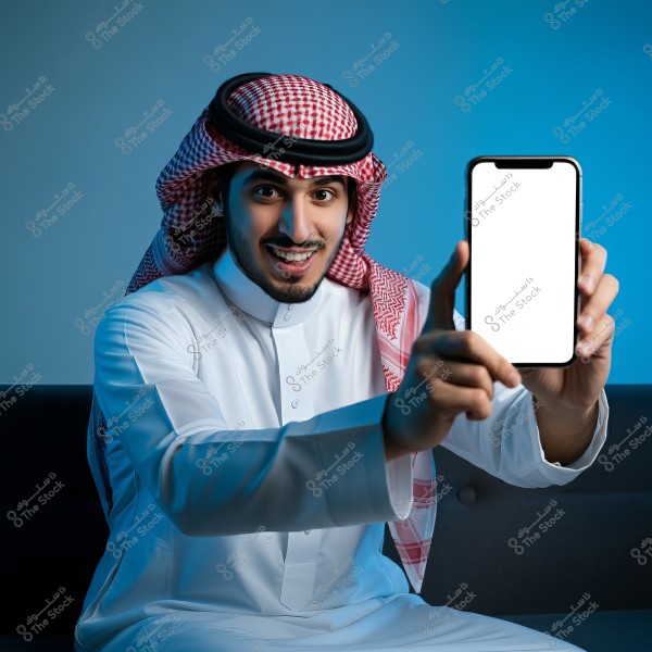 A portrait of a man sitting, wearing a white thobe and a red and white ghutra, holding a smartphone with a blank screen. He is smiling, surrounded by blue lighting.
