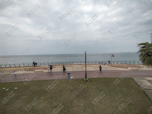 Image shows a promenade along a seafront on a cloudy day. Several people are walking on the pavement, some carrying umbrellas. The sea extends to the horizon in the background. On the left, people are sitting near the metal railing. In the foreground, there is a grassy area with a palm tree in the right corner.