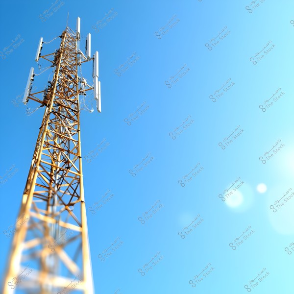 The image shows a tall telecommunications tower with multiple antennas mounted on it, extending into the clear light blue sky. The sunlight illuminates the scene, creating lens flare effects in the image.