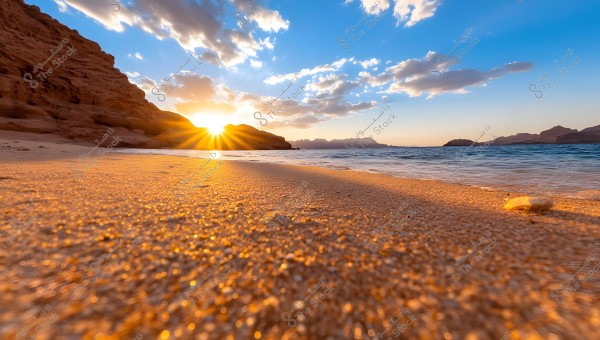 A view of a sandy beach during sunset, with the sun setting between rocky hills on the horizon. The sky is adorned with scattered clouds, and the sea stretches alongside the beach. The golden colors of the sand glisten under the sun\'s rays, creating a picturesque scene.