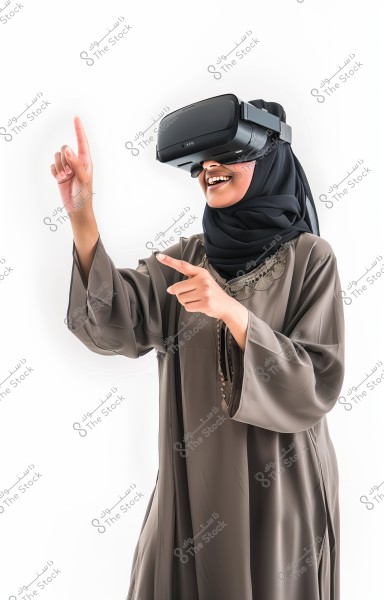 An image of a woman wearing virtual reality goggles, smiling and raising her hand as if interacting with something in the virtual world. She is wearing an abaya and a black hijab, suggesting she may be from Saudi Arabia or another Arab country. The background is white and clean, highlighting the details of the clothing and interaction with technology.