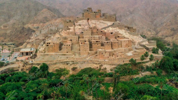 صورة لقرية ذي عين الحجرية في الباحة مبنية على تلة وسط منطقة جبلية. تظهر المباني متراصة ومتراصة بجانب بعضها, وهي مصنوعة من الطوب والحجر. في المقدمة, توجد مجموعة من الأشجار الخضراء والنباتات, مما يضفي تباينًا جميلًا مع الألوان الداكنة للبناء. في الجانب الأيسر, يوجد عدة مباني حديثة ومواقف للسيارات. الخلفية عبارة عن جبال صخرية داكنة تضفي جوًا تاريخيًا.