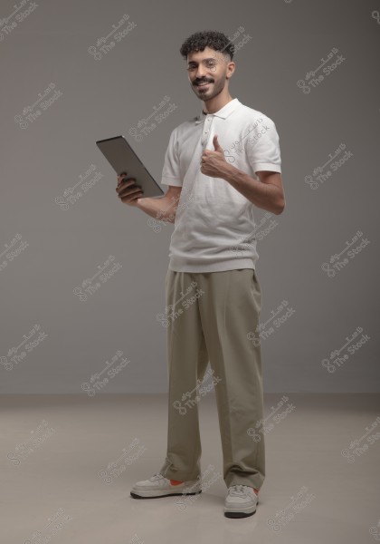 A portrait of a man standing against a gray background, smiling. He is wearing a white polo shirt, beige pants, and sneakers. He is holding a tablet in his right hand and giving a thumbs-up with his left hand. The man has curly hair and a short beard.