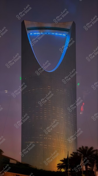 A nighttime image of the Kingdom Tower in Riyadh, Saudi Arabia. The tower is the tallest structure in the image and is characterized by its unique design that resembles a loop at the top. The facade of the tower is illuminated with stunning blue lights while the surrounding sky is dark and starless. Palm trees are seen in the foreground, adding a distinct Arabian atmosphere.