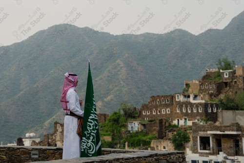 رجل سعودي يرتدي الزي السعودي التقليدي يحمل علم السعودية فوق أحد المباني في قرية رجال ألمع نهارًا بمنطقة عسير، رجل سعودي يحمل علم السعودية احتفالًا باليوم الوطني.